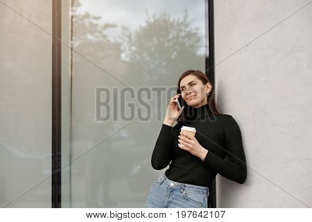 Beautiful student girl in black turtleneck and blue jeans leaning on conceret wall talking on smart phone and holding takeaway coffee in papercup listening to her interlocutor with interest smiling