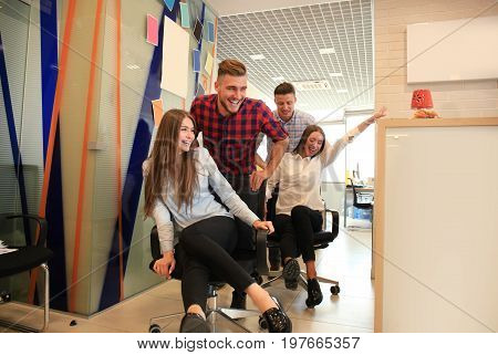 Young cheerful business people in smart casual wear having fun while racing on office chairs and smiling