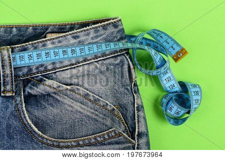 Blue jeans with blue measure tape instead of belt. Upper part of denim trousers isolated on green background. Close up of jeans with measure tape around waist. Healthy lifestyle and dieting concept.
