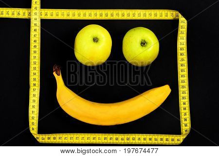 Face made of vegetarian food with happy expression: banana and apples making mouth and eyes framed with yellow measuring tape isolated on black background. Concept of minimalism and happiness