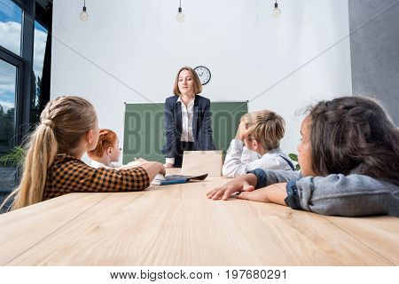 smiling young teacher leaning at desk and looking at cute multi ethnic children in classroom
