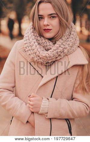 portrait of young beautiful woman walking in stylish warm outfit in sunny autumn day in park wearing pink coat and knitted snood. Casual lifestyle in the city