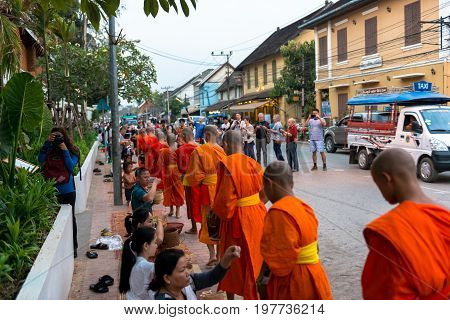 LUANG PRABANG LAOS - MARCH 12 2017: Wide angle picture of local people giving food and money to the monks for the Buddhist Alms Giving Ceremony in Luang Prabang Laos. Tourists take photos.