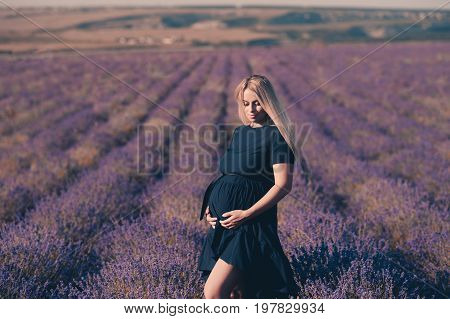 Beautiful blonde pregnant woman standing in lavender field. Wearing stylish dress outdoors. Holding tummy. Motherhood. Maternity.