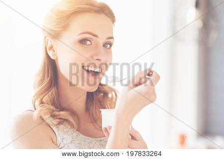 Tasty and healthy. Cheerful beautiful woman holding spoon and eating joghurt while expressing positivity