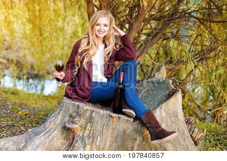 Beautiful woman drinking wine outdoors having picnic in the park. Portrait of young blonde beauty enjoying a glass of red wine sittind on autumn lawn near the lake