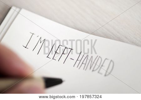 closeup of a young left-handed man handwriting the text I am left-handed in a notepad placed on a table