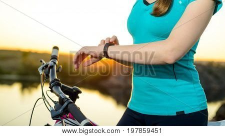 Young Woman Resting with Mountain Bike on the Summer Rocky Trail at Sunset and Using Smartwatch. Closeup of Hands with Smart Watch. Training and Sports Concept.