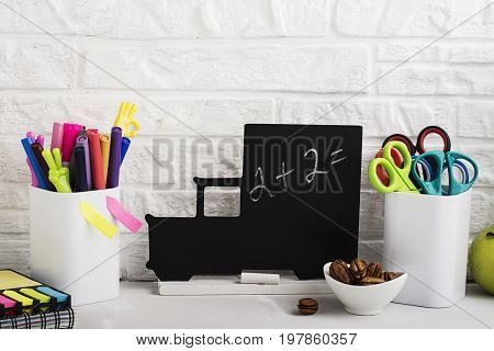 School background: a white brick wall with accessories, pencils, tetrads, scissors, an apple, pecans, a board for a substitute placed in front of her on the table. Selective focus