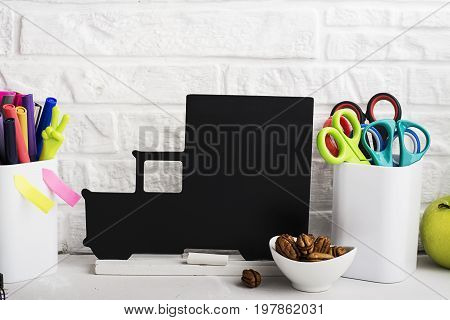 School background: a white brick wall with accessories, pencils, tetrads, scissors, an apple, pecans, a board for a substitute placed in front of her on the table. Selective focus