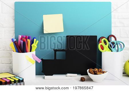 School background: a white brick wall with accessories, pencils, tetrads, scissors, an apple, pecans, a board for a substitute placed in front of her on the table. Selective focus