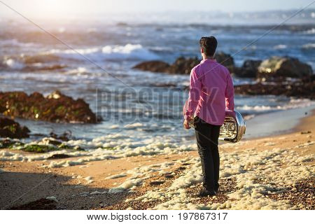 View from the back young musician play trumpet on rocky sea coast during surf. Tuba instrument. 