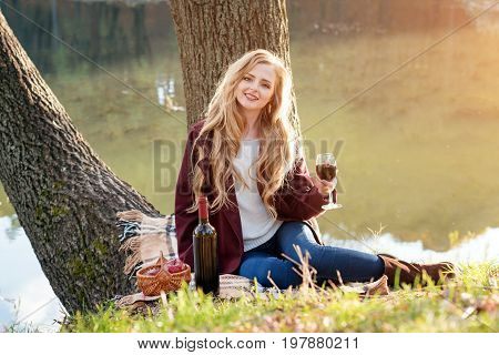 Beautiful woman drinking wine outdoors having picnic in the park. Portrait of young blonde beauty enjoying a glass of red wine sittind on autumn lawn near the lake