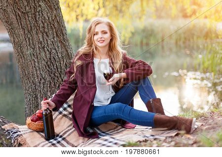 Beautiful woman drinking wine outdoors having picnic in the park. Portrait of young blonde beauty enjoying a glass of red wine sittind on autumn lawn near the lake