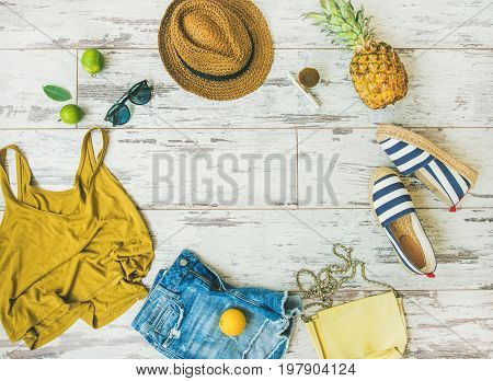 Colorful summer fashion outfit flat-lay. Denim shorts, straw sun hat, yellow top, espadrillas, leather bag, sunglasses, pineapple, limes and lemon over pastel parquet background, top view, copy space