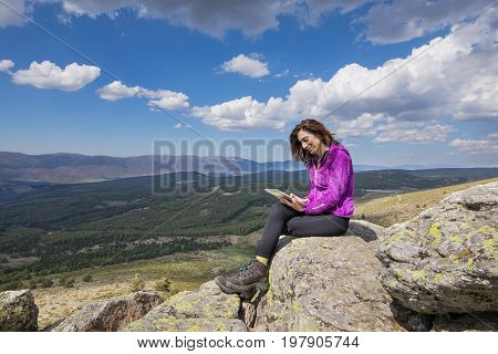 sport hiking or trekking woman with purple jacket sitting on rock peak watching digital tablet next to Lozoya Valley and Guadarrama Park in Madrid Spain