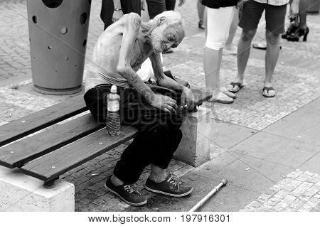 Prague, Czech Republic, August 15, 2015, Editorial photo of skinny homeless beggar who is sitting on bench with indifference other people. Prague, Czech Republic, St. Wenceslaus square