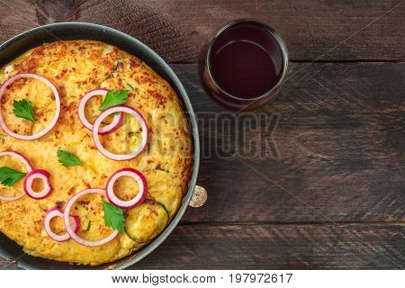 An overhead photo of a Spanish tortilla with potatoes and zucchinis in a tortillera, with a glass of red wine, and a place for text