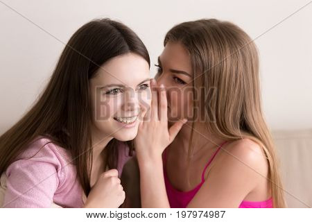 Headshot portrait of two young beautiful teen girls gossiping about boys. Attractive young ladies sharing their secrets, telling scandalous news, whispering in ear rumors and gossips, weave intrigues