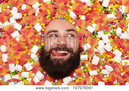 Young bearded man lying in candies and sweets and licking while looking away.