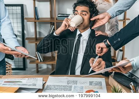 Portrait Of Busy Businessman Drinking Coffee And Sitting At Workplace While Colleagues Helping With