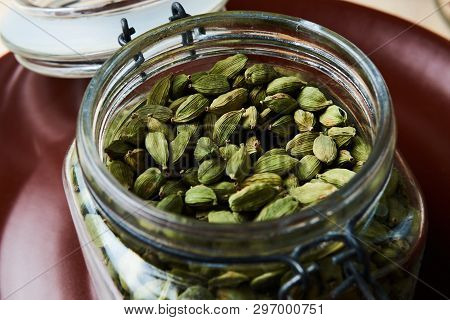 Closeup Proto Of Glass Jar Full Of Green Cardamom Pods On Brown Plate.
