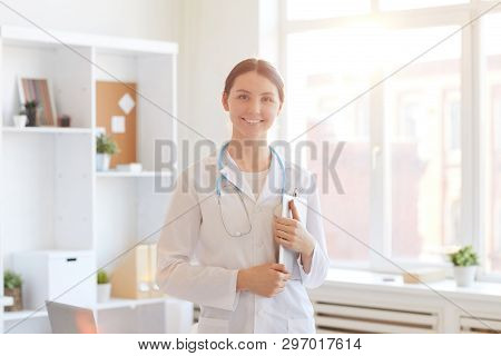 Waist Up Portrait Of Young Female Doctor Smiling At Camera While Posing In Office Lit By Warm Sunlig