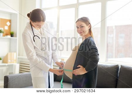 Portrait Of Doctor Measuring Big Pregnant Belly Of Young Woman Smiling At Camera, Copy Space