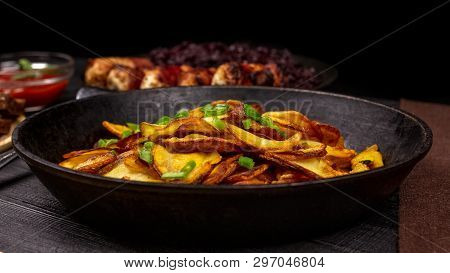 Fried Potato Chips With Greens In A Pot, In Front Of Homemade Sausages And Stewed Cabbage, On A Blac