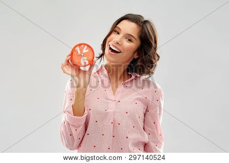 people and bedtime concept - happy young woman in pajama with alarm clock showing half past six over grey background