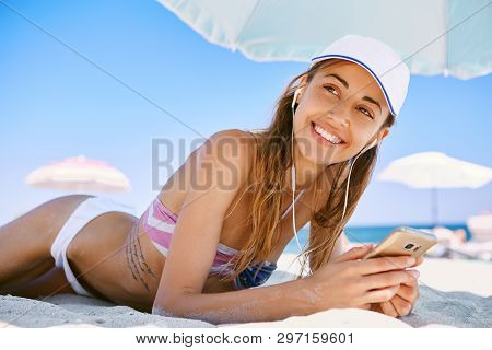 Beautiful Tanned Smiling Woman Is Lying On The Beach On A White Sand, Relaxing And Listen Music From