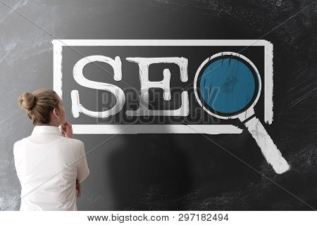 Rear View Of Woman In White Blouse Looking At Term Seo, Search Engine Optimization,  On Chalkboard