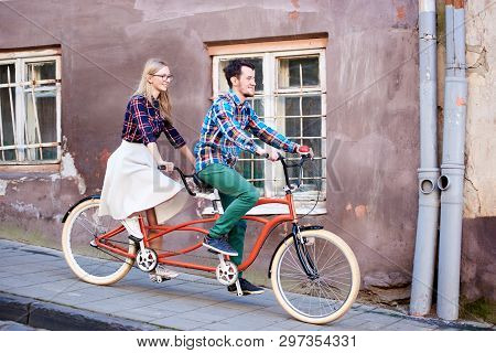 Young Handsome Man And Pretty Blond Woman Riding Tandem Double Bicycle Along Paved Sidewalk On Brigh