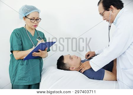 Doctor Litening To Heartbeat Of Young Patient Lying On Couch And Nurse Taking Notes