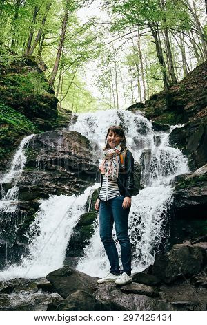 Happy Smiling Tourist Girl In Blue Jeans, Striped Shirt And Black Jacket With Backpack Near To The C