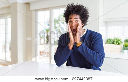 Young african american man wearing casual sweater sitting at home afraid and shocked, surprise and amazed expression with hands on face