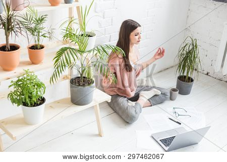 Young Happy Woman Sits On The Floor In A Yoga Pose In A Bright Apartment And Works Behind A Laptop, 