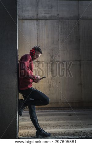 Muscular Athetic Man Wearing Sportswear, Leaning Onto A Concrete Wall, Choosing A Song On A Smart Ph