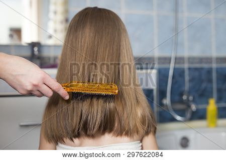 Mother Hand With Brush Combing Long Fair Hair Of Cute Child Girl After Bath On Blurred Interior Back