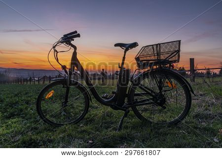 Black Electric Bicycle With Sunset On Light Green Meadow In Spring Color Evening
