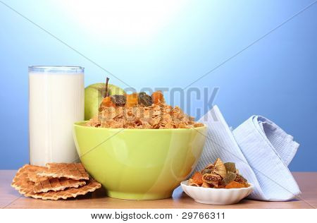 leckere Cornflakes in grüner Schale, Äpfel und Glas Milch auf Holztisch auf blauem Hintergrund
