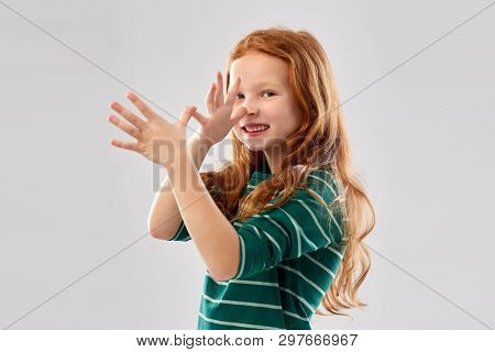 childhood, gesture and people concept - smiling red haired girl in green striped shirt having fun and making big nose over grey background