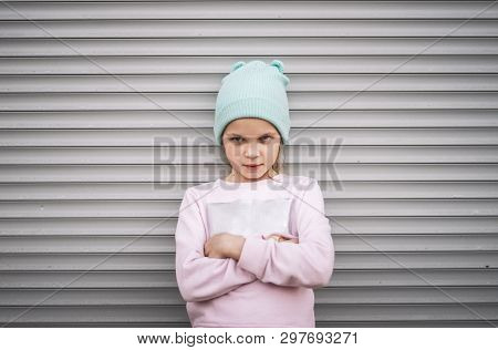 A Serious Child Girl Folded Her Arms Across Her Chest Against The Background Of A City Wall With Gra