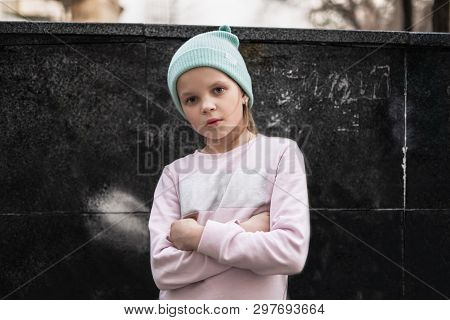 A Serious Child Girl Folded Her Arms Across Her Chest Against The Background Of A City Wall. Street 