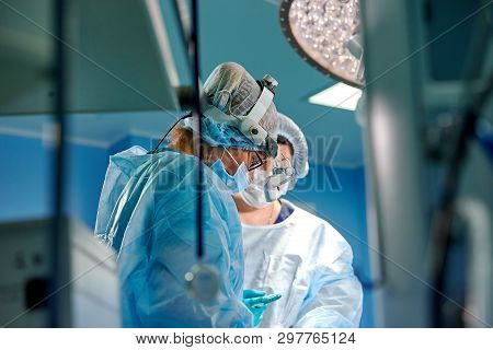 Surgeon Performing Cosmetic Surgery In Hospital Operating Room. Surgeon In Mask Wearing Loupes Durin