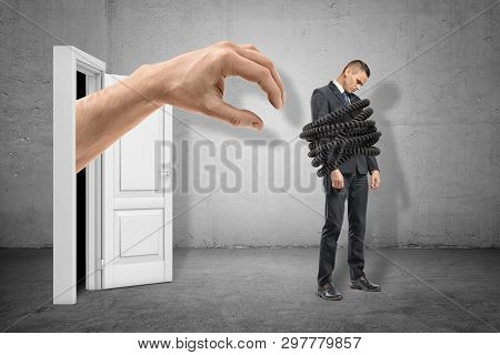 Big Hand Reaching To Young Sad Businessman Tied Up With Telephone Cord On Grey Wall Background