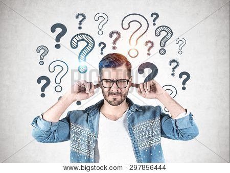 Young Man In Glasses And Casual Clothes Covering His Ears Standing Near Concrete Wall With Question 