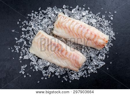 Raw Norwegian skrei cod fish filet as top view on crashed ice on a black board with copy space 