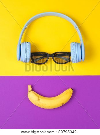 Minimalism Flat Lay Concept. Smiling Face Listens To Music. Headphones, 3d Glasses, Banana On Purple