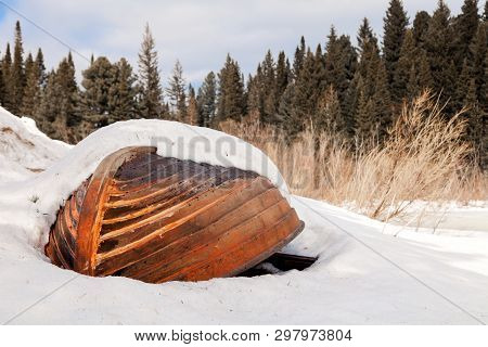 Old Wooden Destroyed Flooded Fishing Boat Lie In Snow On Shore Of Ice-covered Frozen River In Winter